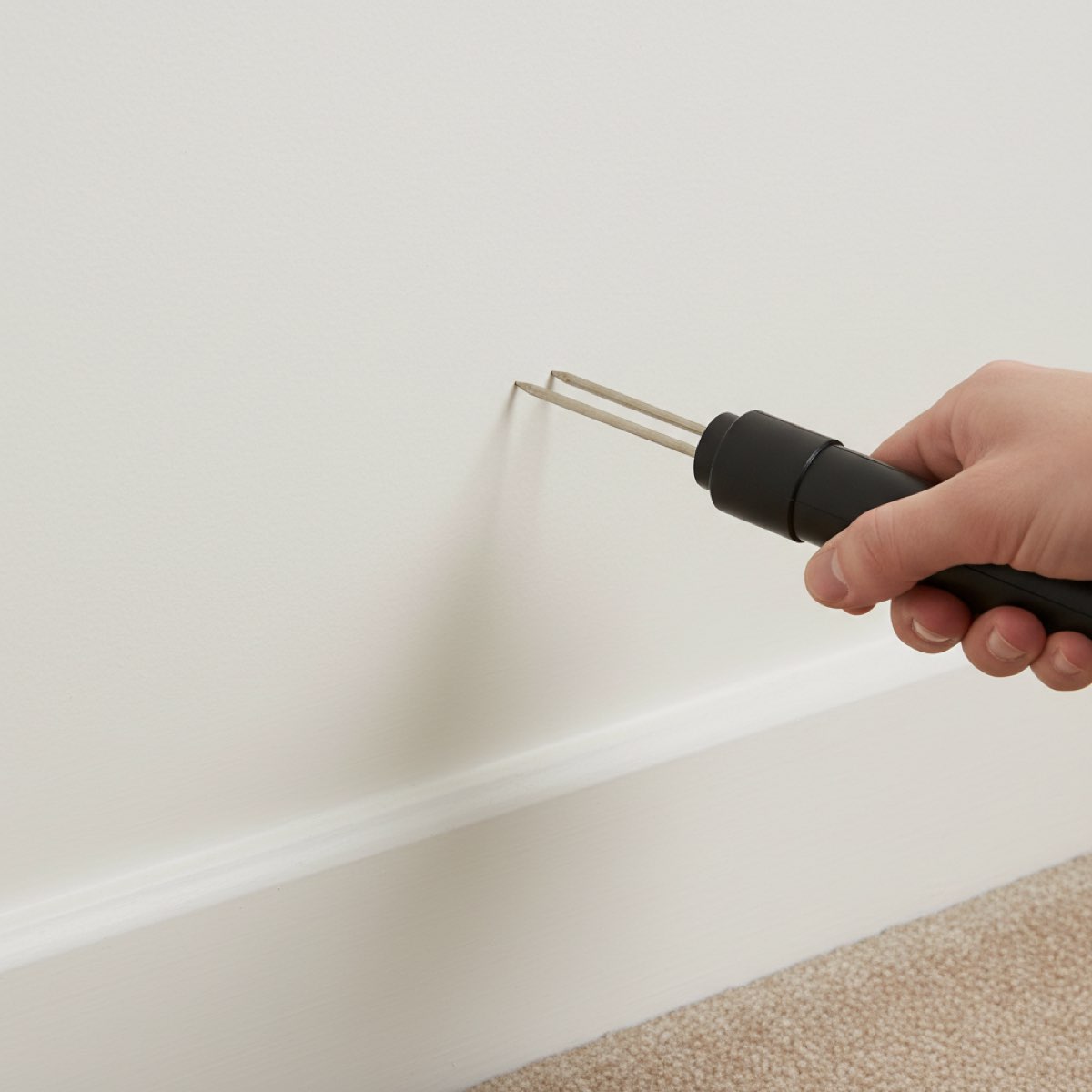 The two metal probe pins of a handheld moisture meter pressed against plain painted plaster just above a skirting board